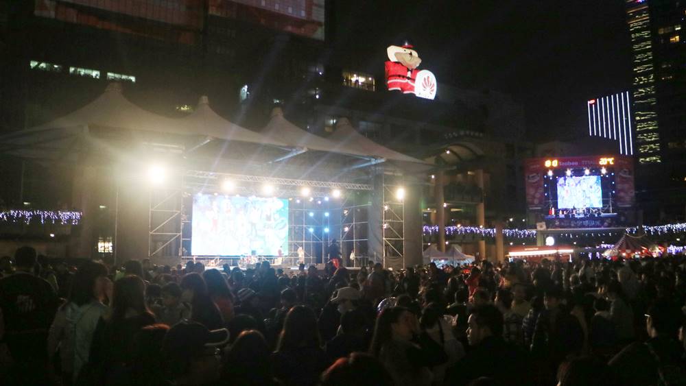 广场上挤满人潮，许多民众坐着边吃边欣赏台上精彩的表演。